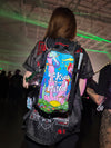 A festival-goer wearing a Lunchbox hydration pack with the "A Kind Mind" skin, featuring a colorful psychedelic "be kind to your mind" design, standing in a warehouse venue with green laser lights and a crowd in the background.