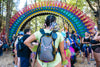 A festival-goer with checkered attire wears a Lunchbox hydration pack with the reflective Disco Queen Skin and floral LED lights, standing before the large, ornate Sherwood Forest archway at Electric Forest.