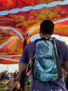 A festival-goer seen from behind wearing a black Lunchbox hydration pack with the "Eternal Serpent" skin, featuring a cosmic blue and green snake with glowing yellow eyes, standing under a vibrant orange and yellow festival canopy.