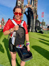 A person at a music festival wearing the Lunchbox Extra Storage Skin 2.0. The black expandable skin is zipped open to reveal a mesh interior pocket holding colorful Kandi bracelets and festival accessories.