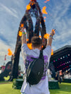 A festival-goer with their back to the camera wearing a black Lunchbox Hydration Sling at a large outdoor music festival. The person has their hands in the air making peace signs, showing how the sling stays secure during movement.