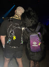 Two festival-goers shown from behind; one wears a black Lunchbox hydration pack with the glittering Midnight Spark Skin, and the other wears a purple and black swirl Insomniac logo skin, both customized with colorful charms and light wires at a night event.