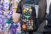 A close-up of a festival-goer wearing a black Lunchbox hydration pack with the "Pin Skin" installed, featuring a dense collection of enamel pins including a pixel-art Calcifer, a Super Mario mushroom, and a "Damascus" brand pin, all secured behind the clear protective shell.
