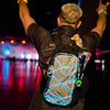 A festival-goer seen from behind with arms raised, wearing a Lunchbox hydration pack with the "Prismatic Skin" and cyan LED light wires, showing the holographic silver surface reflecting vibrant colors under nighttime festival lights.