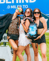 Three women in festival fashion posing together at a music festival; the woman in the center holds a black Lunchbox hydration pack featuring the (Official) Breakaway Skin with its signature blue sky and cloud design.