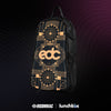 A black Lunchbox hydration pack shown from a side-angle view, featuring the EDC Las Vegas 2025 skin. The skin has a gold Art Deco design with a large "EDC" logo, "All Are Welcome Here" text, and smiley face floral patterns. The pack is set against a dark background with a purple and pink glowing wave