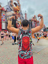 A festival-goer seen from behind, flexing their arms while wearing a black Lunchbox hydration pack featuring the "Ember Bloom" mandala skin, standing in front of a massive, colorful festival stage under an overcast sky.