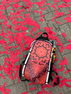 The Ember Bloom Skin zipped onto a black Lunchbox hydration pack, featuring a symmetrical red and orange mandala design with a geometric flower-of-life pattern, shown against a red confetti-covered pavement