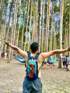 A festival-goer seen from behind with arms outstretched, wearing a Lunchbox hydration pack with the "Geo Jelly" skin featuring a bright orange jellyfish, standing in a forest clearing with hammocks and hanging jellyfish decorations in the trees.