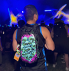 A festival-goer seen from behind wearing a black Lunchbox hydration pack with the "Hexxed" skin, featuring a stack of skulls with glowing neon green eyes, standing in a crowd at a nighttime event with blue stage lights.