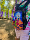 A close-up side view of a festival-goer wearing a black Lunchbox hydration pack with the "Lunar Lasso" skin, featuring a disco cowgirl under a blue moon. The pack is rigged with green and purple elastic bungees and colorful Kandi, shown against a blurred forest background.