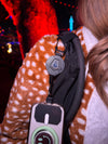 A close-up side view of the Lunchbox Lifeline phone security tether attached to a black backpack strap and a light green smartphone case, featuring the retractable hex-branded metal housing