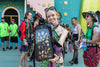 A smiling festival-goer holding a black Lunchbox hydration pack with the "Pin Skin" installed, showcasing a large collection of colorful enamel pins secured behind the clear protective outer shell in a lively outdoor festival setting.